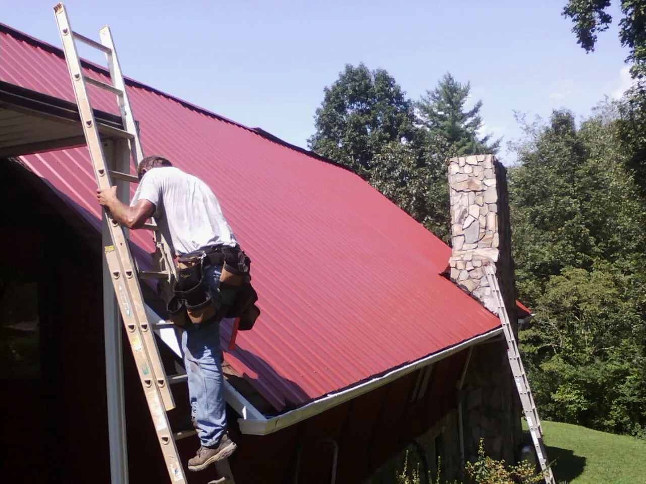Roofer on a ladder beside a red metal roof and stone chimney — Soffit & Fascia Repair by Roofing Brothers in Becket